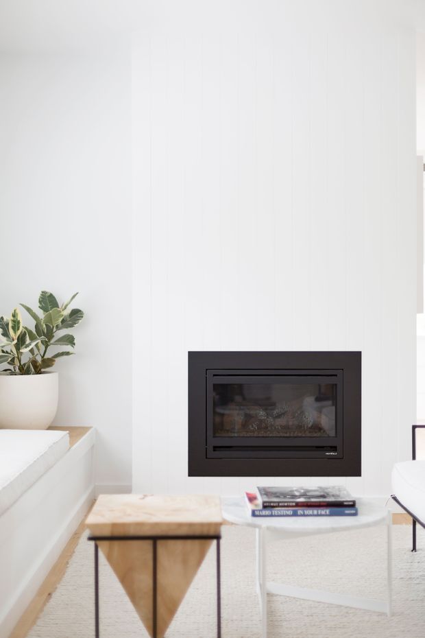 Bright minimalist living room vignette centred on a black built-in fireplace set into a white vertically panelled wall. A white upholstered sofa or built-in bench, potted leafy plant, pale timber coffee table, white side table, stacked books and a light textured rug create a clean, airy interior.