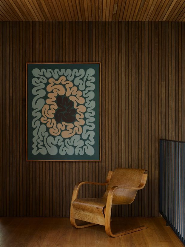 A moody, richly detailed interior corner featuring dark vertical timber wall panelling and a matching timber slatted ceiling. A framed abstract artwork hangs above a sculptural tan upholstered wooden armchair, with timber flooring and a dark metal or timber screen at the right edge. The composition has a warm, refined mid-century character with strong material continuity and low ambient lighting.
