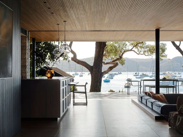 A refined open-plan living and entertaining area with a strong indoor–outdoor connection, overlooking a marina and mountainous waterfront setting. The space features a dark built-in bar or kitchen joinery, expansive glazing, a large sectional sofa, a sculptural central tree, a linear ribbed ceiling, polished concrete flooring, and contemporary pendant lighting.