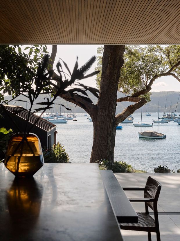 A sheltered waterfront outdoor dining area beneath a timber slatted ceiling, with a long dark stone or concrete dining counter, woven dining chairs, and an amber glass pendant or lantern. Mature trees frame a view across calm water with moored boats and a distant hillside. The scene has a refined coastal atmosphere and strong indoor-outdoor connection.