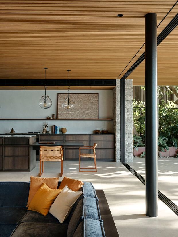 Open-plan contemporary living and kitchen interior with a dark upholstered sofa in the foreground, timber dining chairs, extensive dark timber cabinetry, a long kitchen island, and a large framed artwork. The room has a warm slatted timber ceiling, exposed stone column and wall elements, slim black-framed glazing opening to a planted outdoor area, and a prominent cylindrical structural post. Natural light creates a bright, refined indoor-outdoor atmosphere.