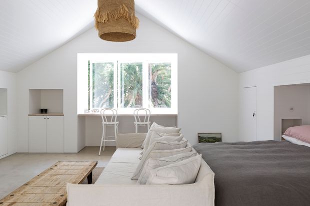 Bright open-plan bedroom with a high pitched ceiling and white painted wall and ceiling finishes. The room combines a neatly made bed with soft neutral grey bedding and a pale upholstered sofa, separated by a rustic timber coffee table. A large window provides views of greenery, while simple white built-in cabinetry, a small desk or vanity area, and woven pendant lighting create a relaxed, minimalist coastal feel.