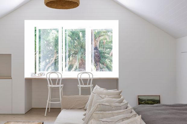 Bright, airy bedroom in a compact gable-roofed space, featuring white horizontal wall panelling, a large three-part window, a built-in window seat or desk with two white chairs, a low bed with layered neutral bedding, and a woven pendant light. Lush planting is visible outside the window.