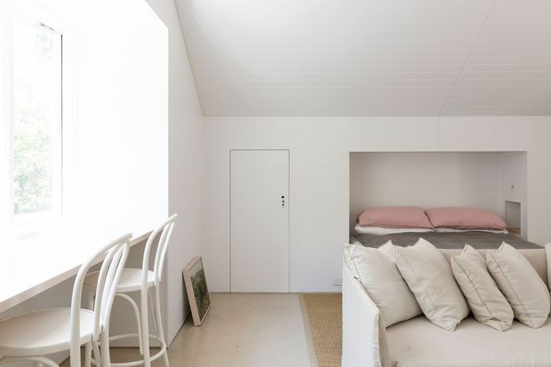 Bright, compact minimalist bedroom with a built-in bed alcove, white bedding and pale pink pillows. A long white desk or dining surface sits beneath a large window on the left with white curved-back chairs, while a simple flush door, pale flooring, and white-painted ceiling and walls create a clean, airy interior.