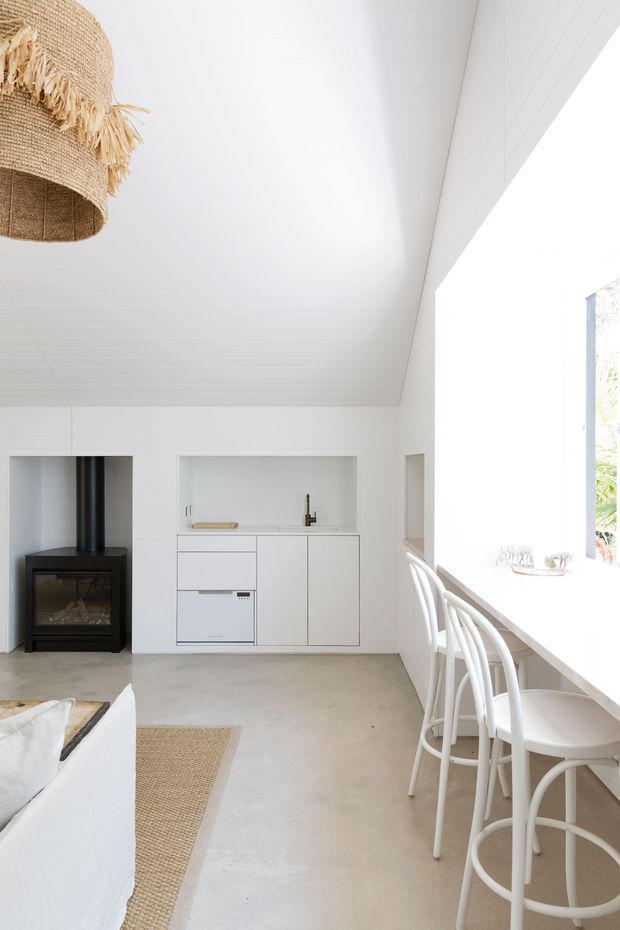 Bright, minimalist open-plan kitchen and living area with white-painted timber wall and ceiling surfaces, a compact white built-in kitchen, pale concrete flooring, and a black freestanding wood-burning stove. A long white breakfast counter runs along large glazing with white bar stools. Natural woven pendant lighting and a jute rug add warmth to the otherwise crisp, airy interior.