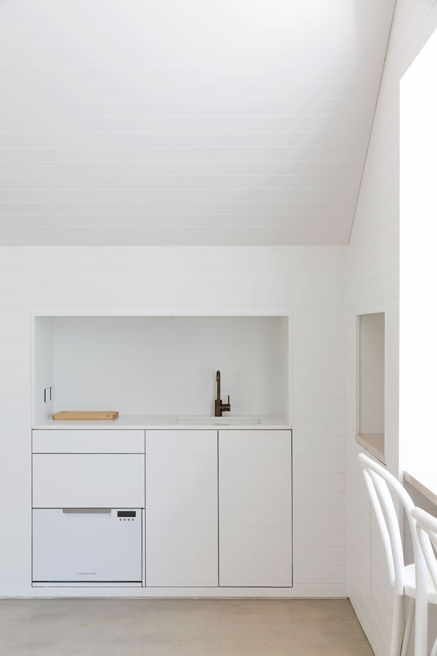 A bright, compact white kitchen or kitchenette with minimalist flat-panel cabinetry, integrated dishwasher, pale benchtop, dark tapware, and a timber chopping board. White horizontal wall and ceiling panelling create a clean, cohesive interior. A small opening and white railing are visible on the right, with a simple concrete-toned floor.