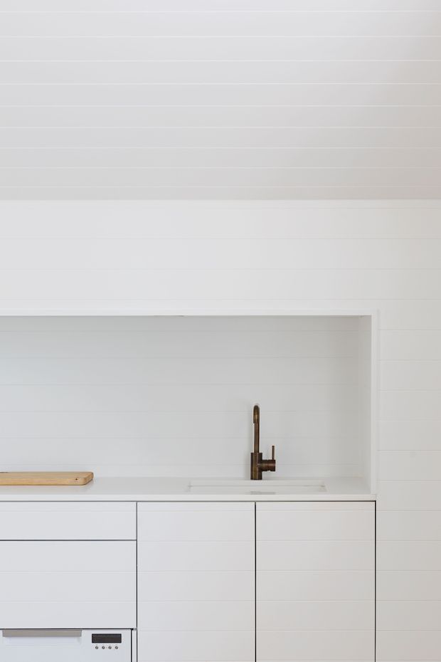 Minimalist white kitchen cabinetry with a pale timber benchtop and integrated sink. The wall and backsplash are clad in horizontal white boards or tiles, with a recessed shelf-like niche above the counter. A slim bronze or dark metal gooseneck tap provides the main visual accent, while integrated appliances are visible below.