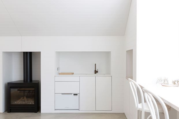 Bright minimalist open-plan kitchen and dining area with white built-in cabinetry, a concealed appliance, pale flooring, a recessed sink niche, and a black freestanding wood-burning stove. A white dining counter with curved white chairs is visible at right. The room has a clean, light-filled contemporary interior with subtle horizontal ceiling panelling.