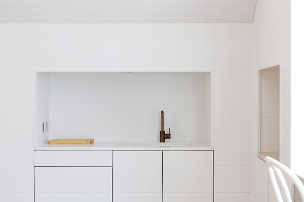 Minimalist white kitchen or kitchenette with a compact built-in cabinet run, integrated sink, bronze-toned gooseneck tap, white horizontal wall panelling, and a recessed open shelf above the benchtop. A small timber cutting board adds warmth, while partial white bar stools are visible at the right edge.