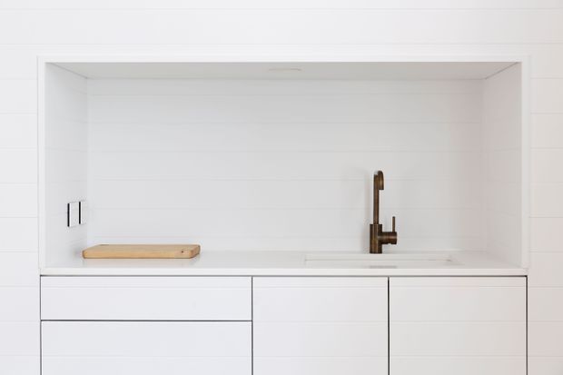 Minimalist white kitchen or kitchenette joinery with a long recessed open shelf, integrated pale timber work surface, white cabinetry, and a compact sink with a dark metal tap. The image is tightly framed on the fixed cabinetry and benchtop, with clean horizontal lines and very limited decoration.