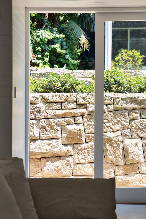Interior view looking through large floor-to-ceiling glazed doors or windows toward a landscaped garden and textured sandstone retaining wall. The room foreground includes part of a neutral upholstered sofa, while the glazing has slim white frames and is set within white horizontal wall and ceiling panelling. Bright natural light and lush planting create a calm indoor-outdoor connection.