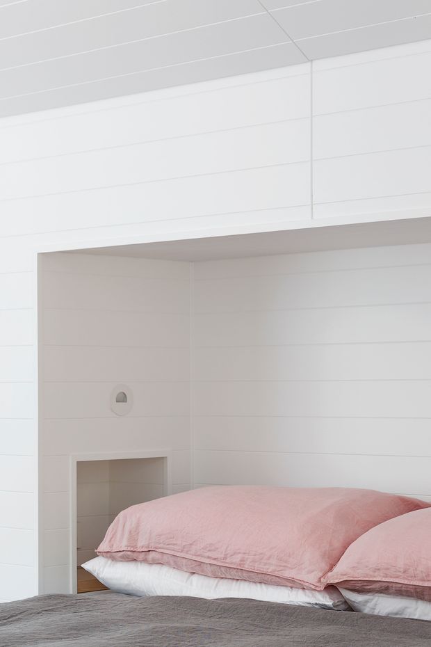 A close-up view of a compact bedroom with a low bed in the foreground, layered white and blush-pink bedding, and a grey bedcover. The room has white horizontal wall panelling, a white paneled ceiling, and a recessed wall niche or opening beside the bed. The overall appearance is bright, restrained, and contemporary with soft pastel accents.