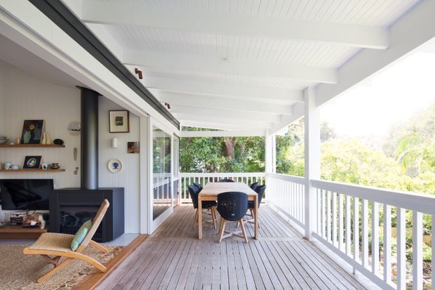 Bright covered timber veranda or deck adjoining an open-plan living area. The space features white painted posts, balustrades and a beadboard-style ceiling, with a timber dining table and chairs overlooking leafy surroundings. Sliding glass doors connect the deck to the interior, where a fireplace, shelving and lounge seating are visible.