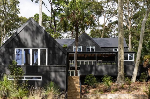 Exterior view of a dark-clad, multi-wing house set among mature trees and native landscaping. The building has steep gabled roof forms, black vertical cladding, white-framed windows, a raised timber entry stair, and a covered upper-level outdoor balcony or verandah with slim metal balustrading.