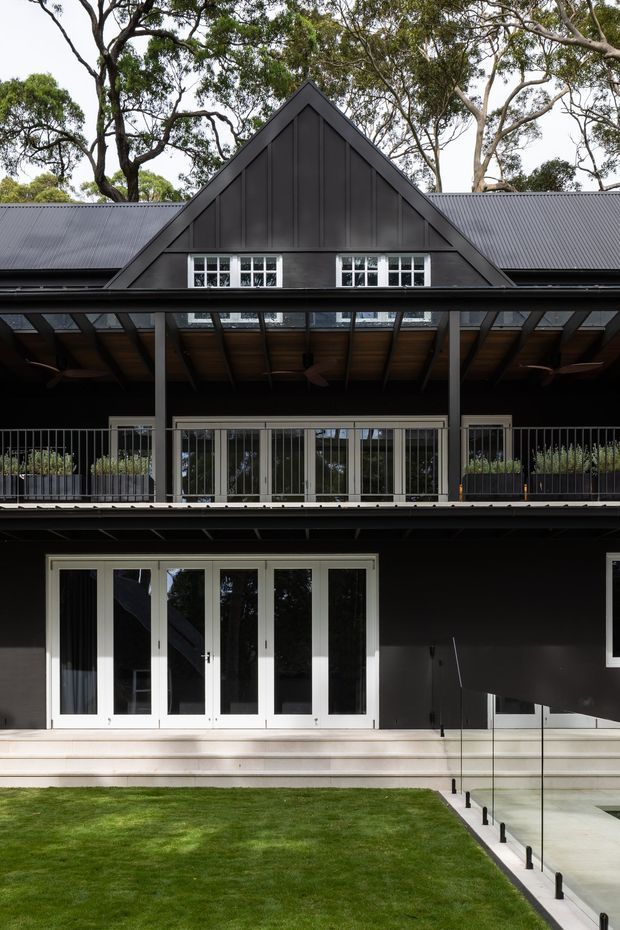 Front exterior view of a substantial dark-painted, two-storey house set among mature trees. The facade features a prominent central gable with vertical cladding, a pitched corrugated metal roof, white-framed multi-pane windows and French doors, a covered upper-level terrace with glass balustrades, and a lower terrace accessed by broad steps. A neat lawn and clear glass pool or terrace barrier occupy the foreground.