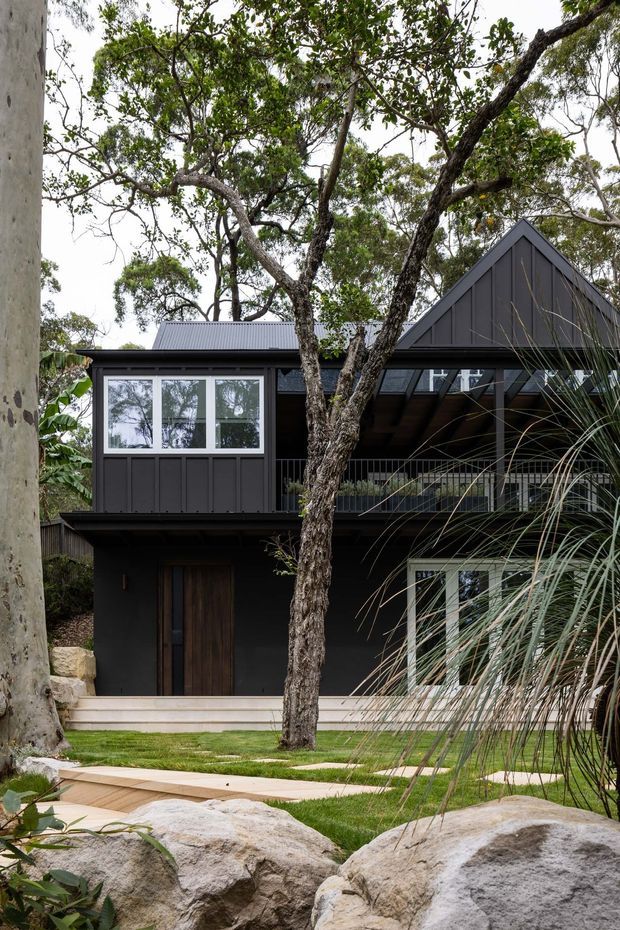 Exterior view of a dark-clad, multi-level house set among mature trees and large natural rocks. The building has a steep gabled roof, corrugated metal roofing, vertical dark cladding, extensive white-framed glazing, a covered upper-level balcony or walkway, and a landscaped garden path in the foreground.