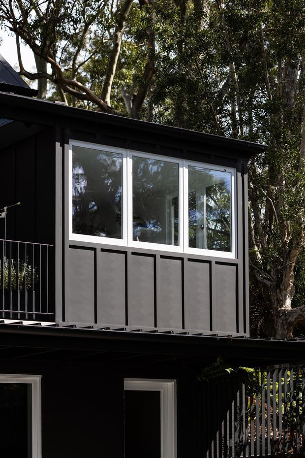 Exterior view of a compact, dark-clad multi-level building set among dense mature trees. The facade features black vertical and panelled cladding, white-framed windows, a simple low-pitched roof, and a narrow upper-level balcony with metal railings. Reflections in the glazing reveal surrounding vegetation and interior glimpses.