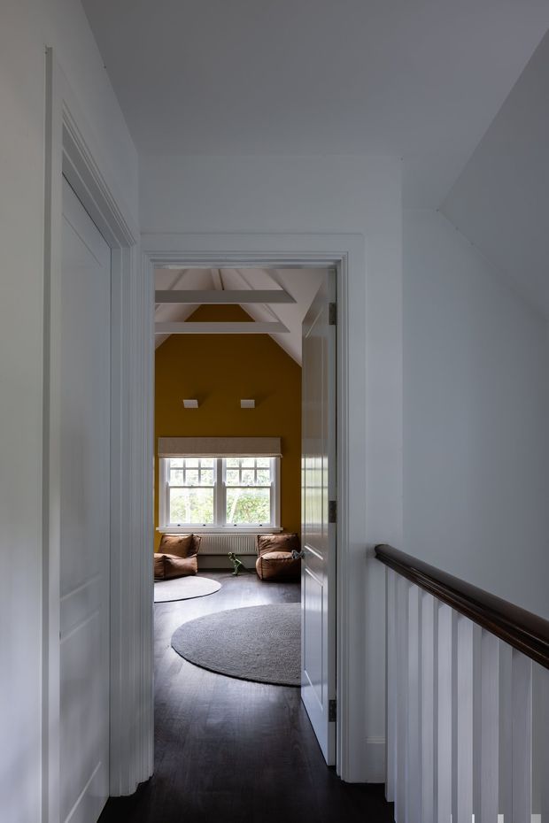 A narrow, white-painted upstairs hallway and landing with dark timber flooring and a dark wood handrail, looking through an open paneled door into a cozy sitting room. The sitting room has mustard-yellow walls, a pitched ceiling with exposed white-painted structural beams, a large multi-pane window, brown leather seating, and round grey rugs.