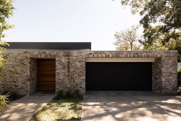 Single-storey contemporary residential facade with a low, flat roofline and extensive exposed brickwork. The view focuses on a wide black garage door beside a recessed timber entry door, with concrete paving and subtle landscaping in front. Mature trees frame the property, and strong sunlight creates dappled shadows across the driveway and facade.