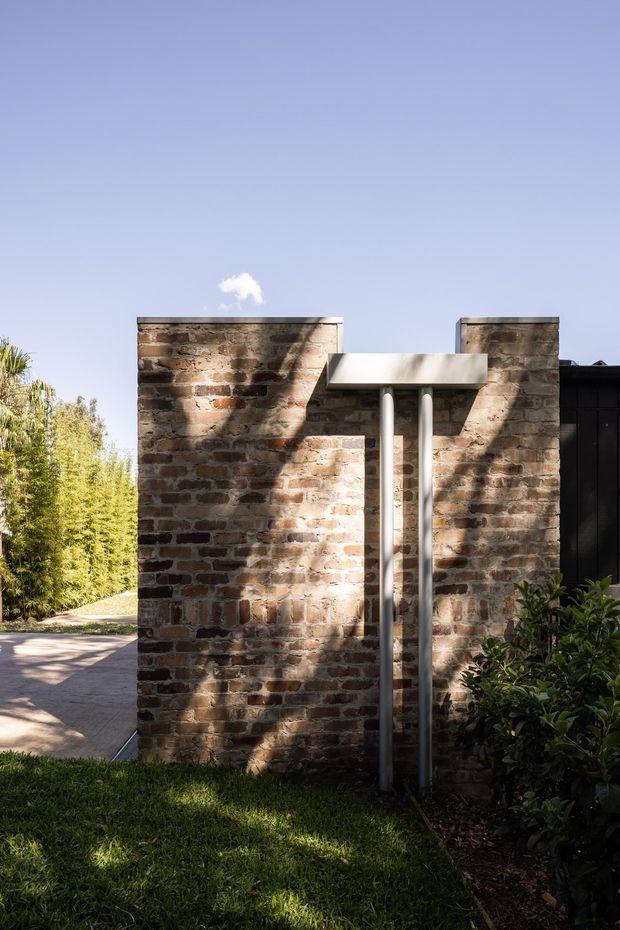 Exterior architectural detail viewed from a landscaped garden path. A tall, textured exposed-brick wall forms a courtyard or entry feature, with a minimalist projecting pale concrete canopy supported by two slender metal posts. Dark cladding is partially visible to the right, while planted borders, lawn, paving, and strong tree shadows surround the structure under a clear blue sky.