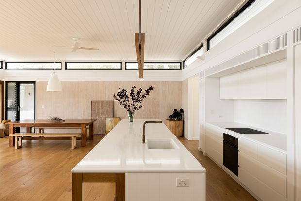 Bright open-plan kitchen and dining area with a large white island, integrated sink and dark tapware, white built-in cabinetry, timber flooring, a timber dining table with benches, and a light timber-lined ceiling. High-level clerestory windows and extensive glazing provide natural light, while a concealed rangehood and induction cooktop form part of the minimalist kitchen wall.