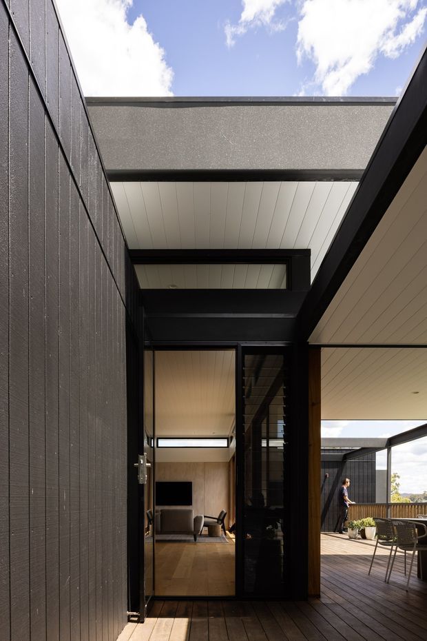 A contemporary residential courtyard or terrace passage framed by dark vertical cladding and black steel-framed glazing. The view looks through an open glazed connection into a warm timber-floored living room, while a covered outdoor deck with dining furniture occupies the right side. White-lined soffits, strong geometric roof beams, and an open skylit void create a crisp indoor-outdoor composition.