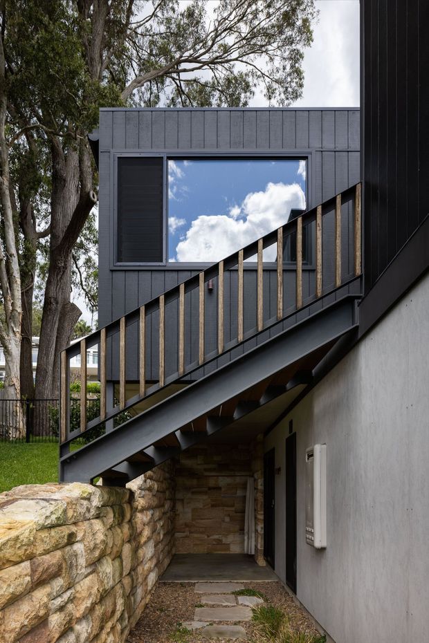 Exterior architectural photograph of a modern multi-level residence set among mature trees. A dark metal staircase with timber-toned vertical balusters rises diagonally beside a charcoal vertical-panel-clad upper volume, with a large reflective window. The lower level has smooth light-grey walls, a rugged natural-stone retaining wall, and a narrow paved walkway beneath the stair.