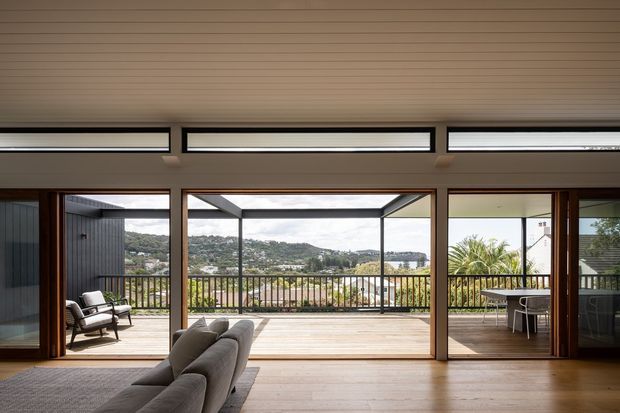 A bright contemporary living room opening onto a large timber deck through extensive floor-to-ceiling glazing. The room has a linear timber-lined ceiling, warm timber flooring, slim dark-framed sliding doors, and a grey sectional sofa. Beyond the deck is a balustraded outdoor terrace with lounge and dining furniture, overlooking a green hilly suburban landscape.