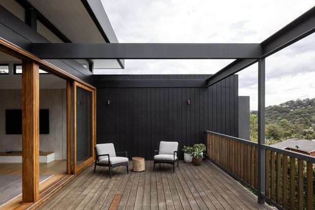 A contemporary elevated timber deck terrace adjoining a modern home. The covered outdoor area has exposed dark steel beams, warm timber-framed glazing and sliding doors, a dark vertical-panel feature wall, timber decking, and a timber balustrade. Two lounge chairs and potted plants create a compact outdoor seating area, with a leafy hillside visible beyond the railing.