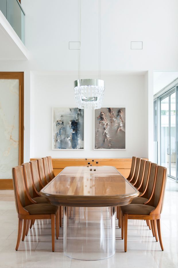Bright, refined dining room with a long polished dining table, a transparent cylindrical pedestal base, and matching wood-framed upholstered dining chairs. White walls and polished pale flooring create an airy setting, with a recessed timber sideboard, abstract artwork, a sculptural pendant light, and extensive glazing visible to the right. A partial upper-level glass balustrade is visible at the left.