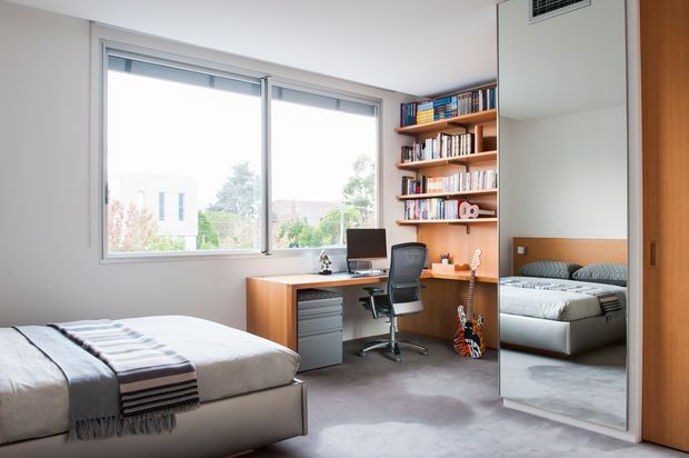 Bright contemporary bedroom with a built-in study nook, timber desk, office chair, extensive open shelving filled with books, and a large full-height mirrored wardrobe door reflecting the bed. The room has pale walls, grey carpet, generous glazing, and a minimal neutral palette with warm timber accents.