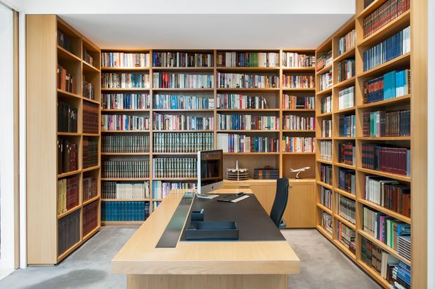 A compact, floor-to-ceiling library and study lined with extensive warm timber shelving filled with books. A central light timber desk with a dark work surface and office chair occupies the room, with pale concrete flooring and white ceiling and wall surfaces. The space is bright, orderly, and highly functional.