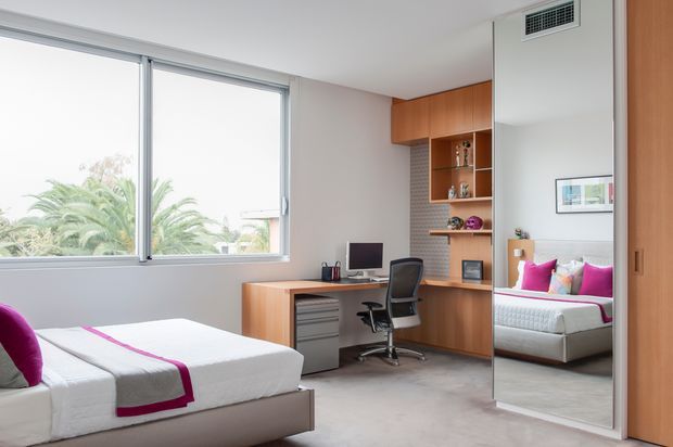 Bright contemporary bedroom with a built-in timber study nook and shelving, an office chair and computer desk, large windows, carpeted flooring, and a full-height mirrored wardrobe door reflecting the bed and colourful cushions.