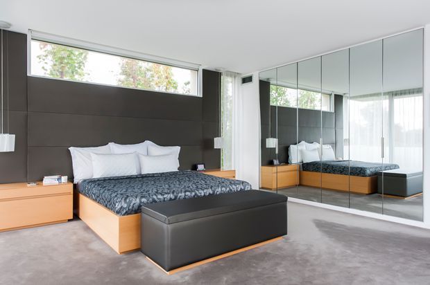 A contemporary bedroom with a low-profile bed, dark upholstered headboard wall, light timber bedside cabinetry, mirrored built-in wardrobe doors, and a matching upholstered bench at the foot of the bed. The room has a soft grey carpet, white walls and ceiling, sheer curtains, and generous natural light.