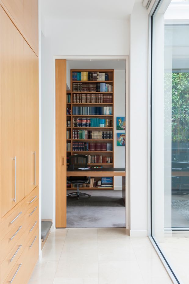 Bright modern hallway with full-height light timber built-in cabinetry and drawers on the left, large floor-to-ceiling glazing on the right, and a timber sliding door opening to a compact home office. The office contains a wall-to-wall bookcase filled with books, a built-in desk, and an office chair. Pale tiled flooring and white walls create a clean, airy interior.