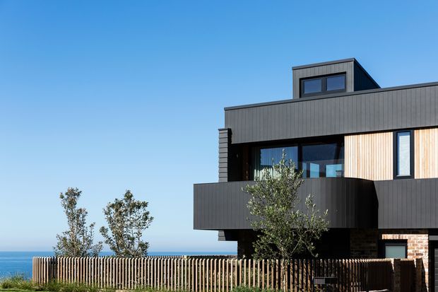 Exterior view of a contemporary multi-level coastal residence with a strongly horizontal composition. The dark vertical-clad facade includes projecting balcony forms, large glazed openings, pale vertical timber screening, a rooftop upper volume, and a small section of exposed brickwork. A timber picket fence and planted trees sit in front, with the ocean visible beyond.