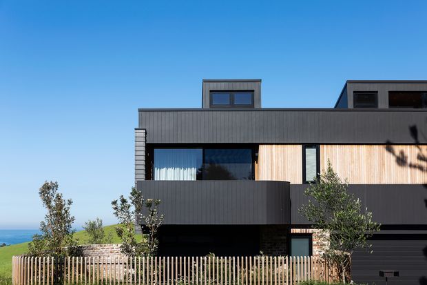 Modern multi-storey residential facade with dark vertical cladding, warm timber wall sections, expansive glazing, projecting balconies, rooftop structures, and a vertical timber slat fence. The house sits on a coastal-looking landscaped site under a clear blue sky.