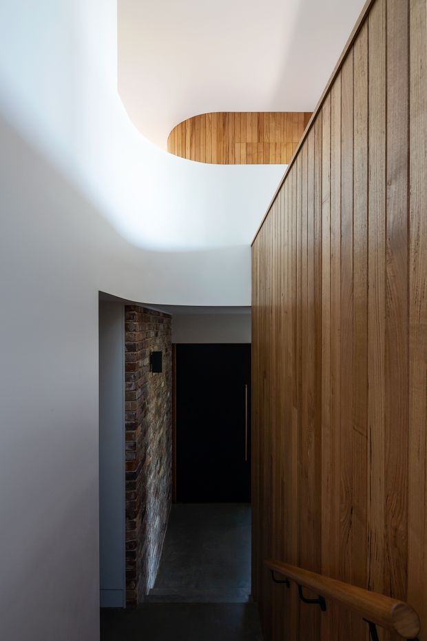A sculptural interior hallway or stair landing with smooth curved white plaster walls and ceiling, a warm vertical timber-panelled wall with integrated handrail, and an exposed brick wall leading toward a dark doorway. The composition emphasises contrasting natural timber, brick, dark flooring, and dramatic indirect daylight.