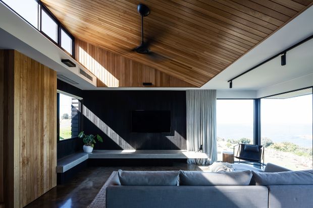 A modern open-plan living room with a large grey sectional sofa facing a wall-mounted television and dark built-in media joinery. The room features extensive warm timber wall and ceiling lining, polished dark flooring, high clerestory glazing, broad windows, white walls, black track lighting, and a ceiling fan. Bright coastal or landscape views are visible through the glazing.