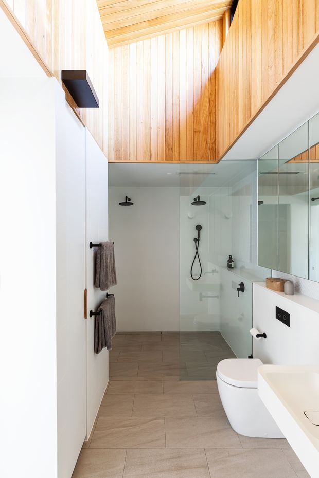 Bright contemporary bathroom with a walk-in glass shower, wall-mounted toilet, white vanity, mirrored cabinetry, and warm vertical timber lining across the sloped ceiling and upper walls. Neutral grey-beige tiled flooring, white surfaces, and matte black plumbing and hardware create a clean, refined interior.