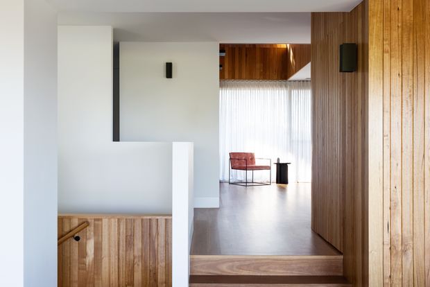 Interior view of a modern timber-lined hallway or landing, looking toward a bright sitting area. A short set of timber steps leads up to an open room with a red lounge chair and small black side table. White plaster walls contrast with extensive vertical timber wall panelling and warm timber flooring, while sheer curtains diffuse daylight. A stair opening with timber balustrade or cabinetry is visible in the foreground.