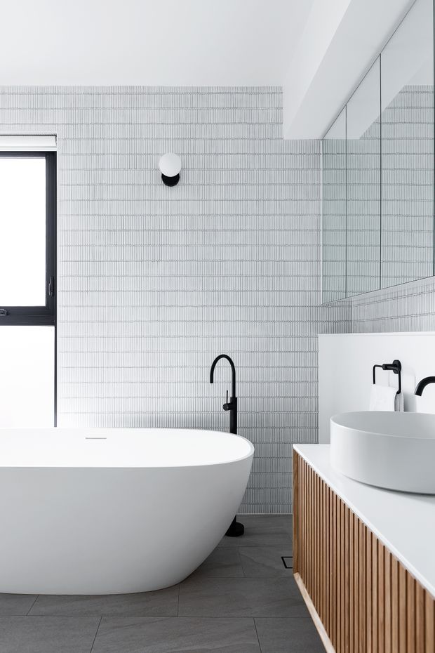 A bright contemporary bathroom featuring a freestanding white bathtub, round vessel basin, black tapware, a timber slatted vanity, large mirrored cabinetry, and fine vertically textured grey wall tiles. Dark grey floor tiles and a black-framed window add contrast to the otherwise restrained palette.