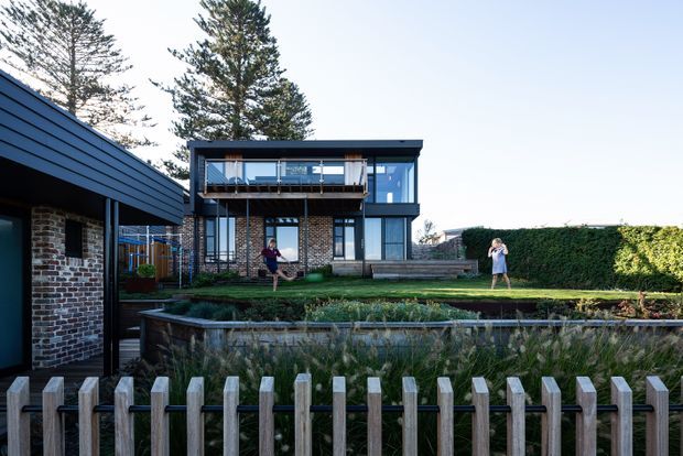 Exterior view of a contemporary multi-level residence with dark horizontal cladding, exposed brick walls, extensive glazing, and a balcony with a glass balustrade. The house is set behind a timber picket fence and landscaped raised garden beds, with mature trees and hedging around the property.