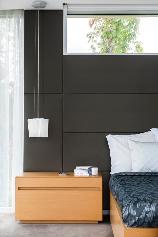 A contemporary bedroom detail featuring a dark charcoal panelled feature wall, a narrow horizontal window, sheer curtains, a suspended white pendant light, and light timber bedside cabinetry and bed framing. The bed is dressed in white pillows and dark patterned bedding, with a soft neutral carpet underfoot.