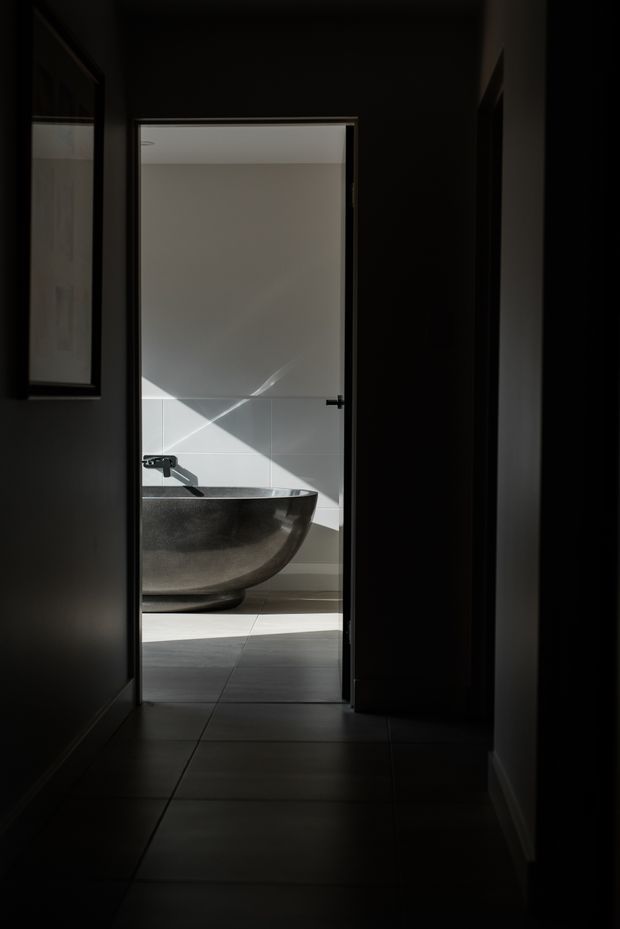 A dramatic, low-light interior view down a dark tiled hallway toward an open bathroom. The bathroom contains a sculptural dark freestanding bathtub, wall-mounted tapware, pale tiled walls, and strong diagonal sunlight creating geometric shadows. Dark door and window frames contrast with the bright bathing space.