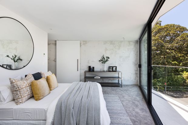 Bright contemporary bedroom with a low-profile upholstered bed dressed in white linen, textured neutral cushions, and a grey knitted throw. The room features a large round mirror, a slim black console table, a concealed white door, pale mottled feature walls, grey carpeting, and expansive black-framed glazing opening to leafy surroundings and a balcony or terrace.