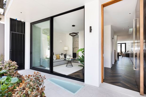 A contemporary residential courtyard viewed from outdoors, with a small linear pool in the foreground, landscaped planting, black-framed full-height glazing, and a covered entry. Through the glazing is a refined living room with a grey sofa, light rug, sculptural pendant, and indoor plants. The open timber-framed door leads into a hallway with dark timber flooring and further living spaces beyond.