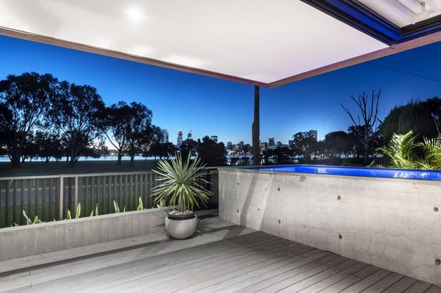 A covered outdoor terrace overlooking a waterfront or lakeside setting at dusk. The space features grey timber decking, a concrete privacy wall with an integrated illuminated blue-edged pool or water feature, vertical balustrading, a structural post, and potted planting. The open roofline frames views toward trees and a distant urban skyline.