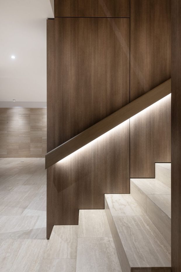 Contemporary interior stairway with broad pale stone or porcelain-tiled steps, dark timber wall panelling and integrated warm LED strip lighting beneath the diagonal handrail. The adjacent open floor area continues the light stone finish, creating a refined, minimalist transition space.
