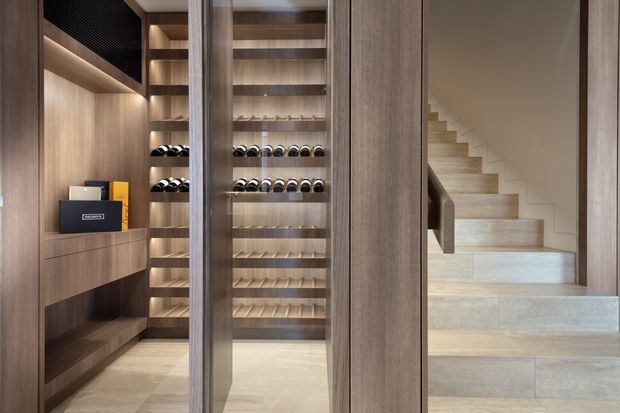 A refined built-in wine storage and display area beside a light stone staircase. The space features extensive warm timber cabinetry, open shelving, bottle racks, integrated lighting, a glass-fronted display cabinet, and pale stone flooring and stair treads. The design is clean-lined and contemporary with a warm, luxurious atmosphere.